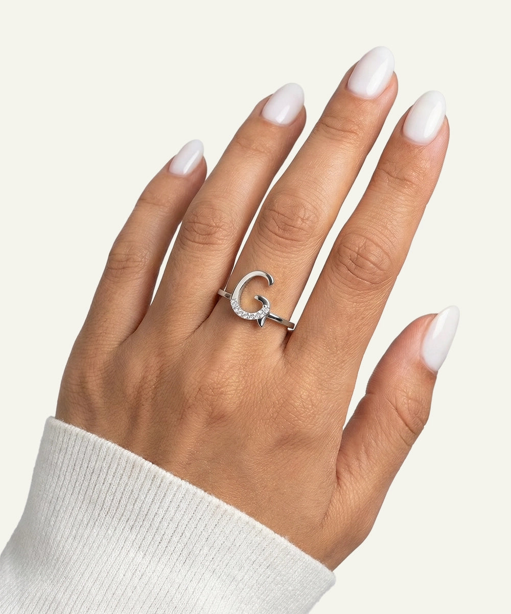 Hand wearing a silver ring with a 'G' design on a light background