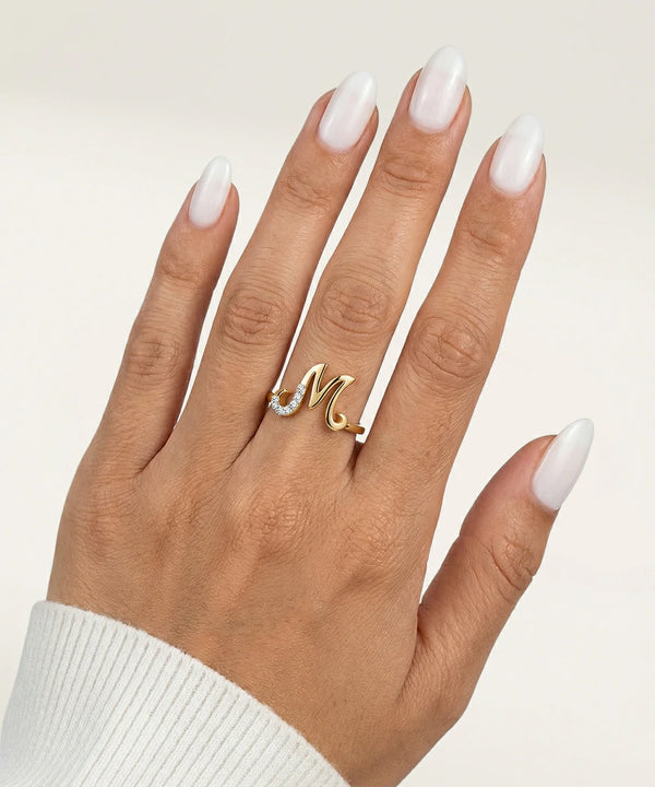 Hand wearing a gold ring with a letter 'M' on a neutral background