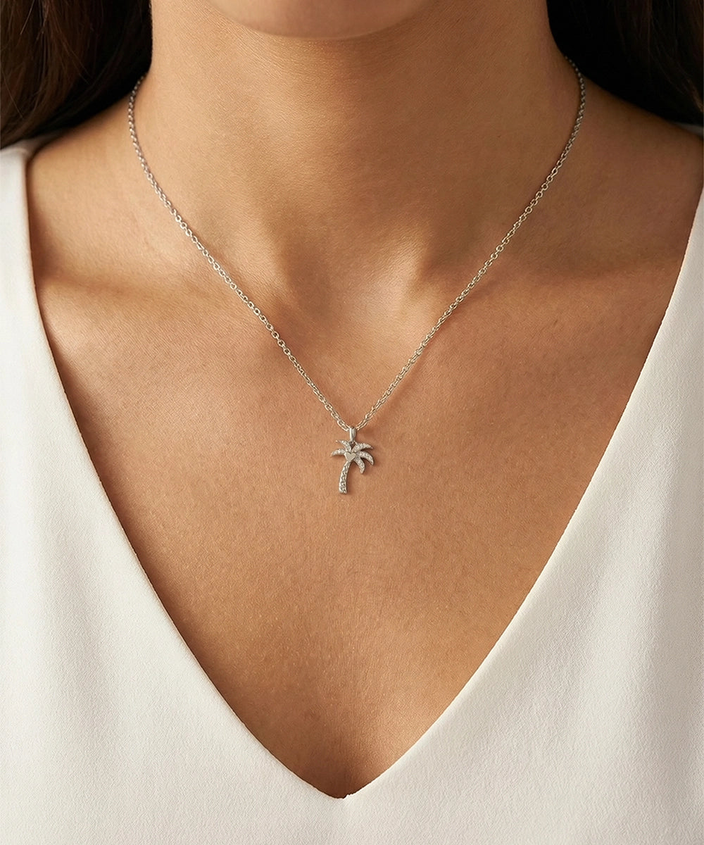 Silver necklace with a palm tree pendant worn by a person in a white top.