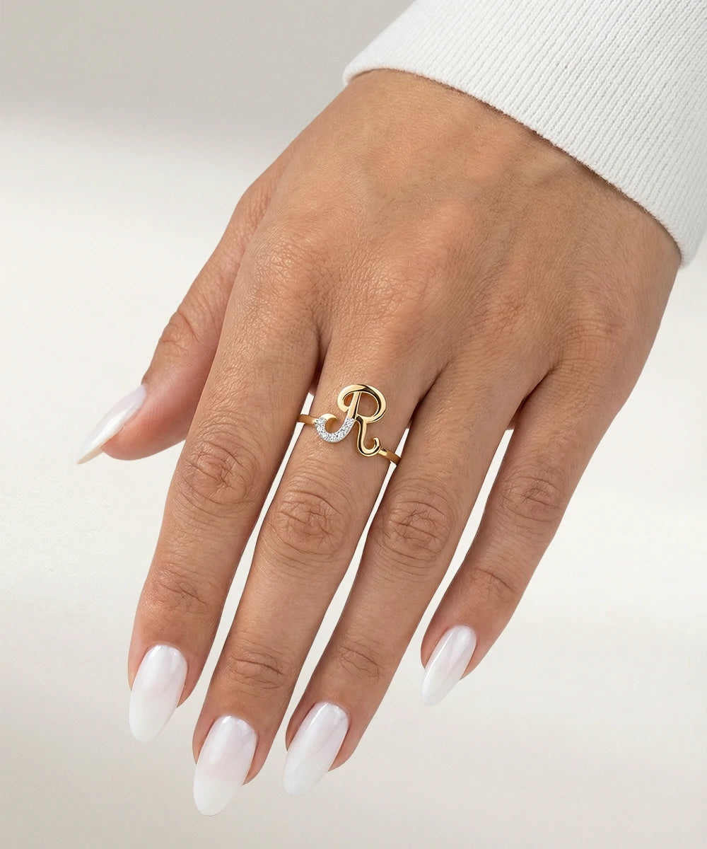 Hand wearing a gold ring with a 'R' on a light background