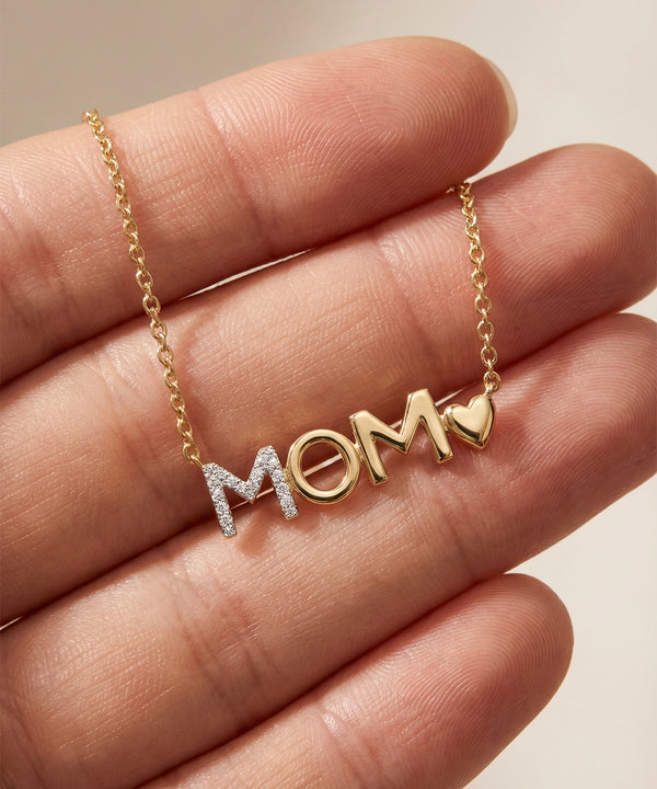 Gold necklace with 'MOM' pendant held between fingers on a neutral background
