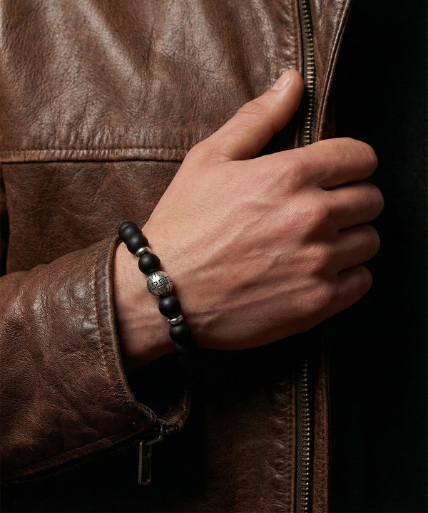 Hand wearing a black beaded bracelet on a brown leather jacket.