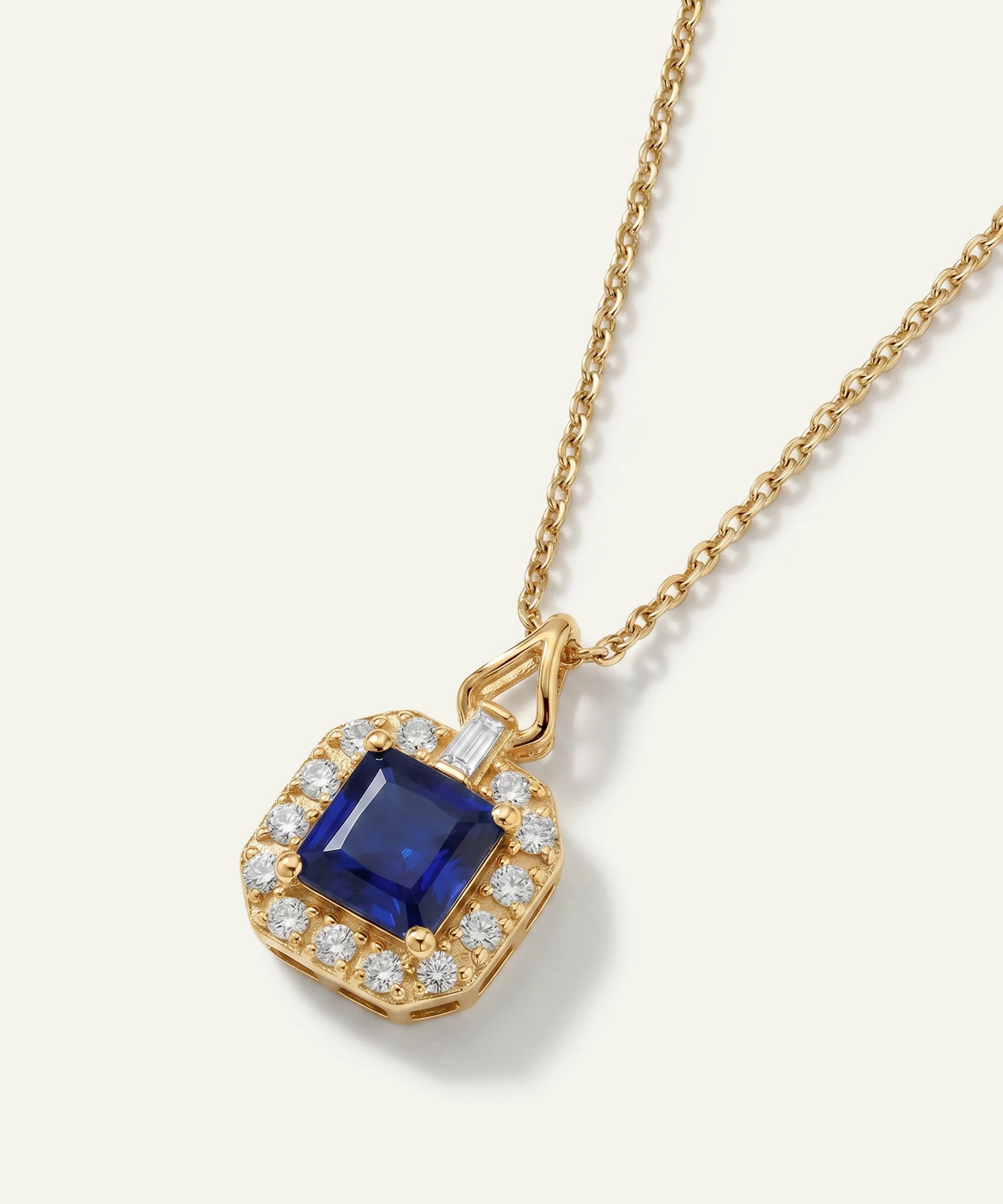 Elegant yellow gold pendant necklace featuring a cushion-shaped halo of round diamonds surrounding a deep blue emerald-cut sapphire, suspended from a delicate gold chain on a light neutral background.