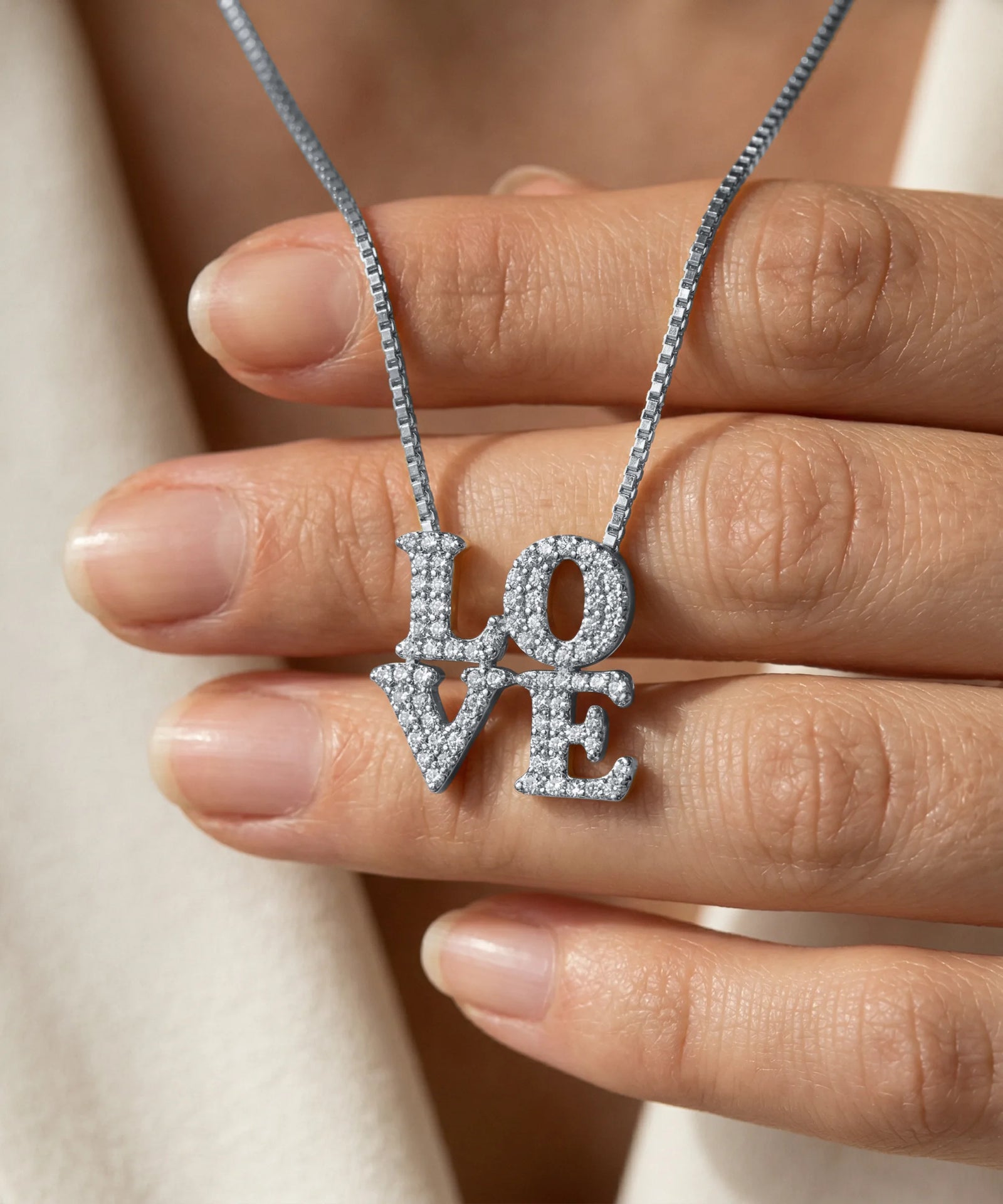 Silver 'LOVE' necklace held between fingers against a neutral background