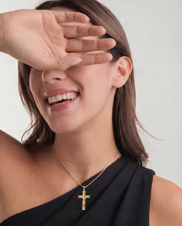 Woman wearing a gold necklace with a cross pendant, covering her eyes with her hand.