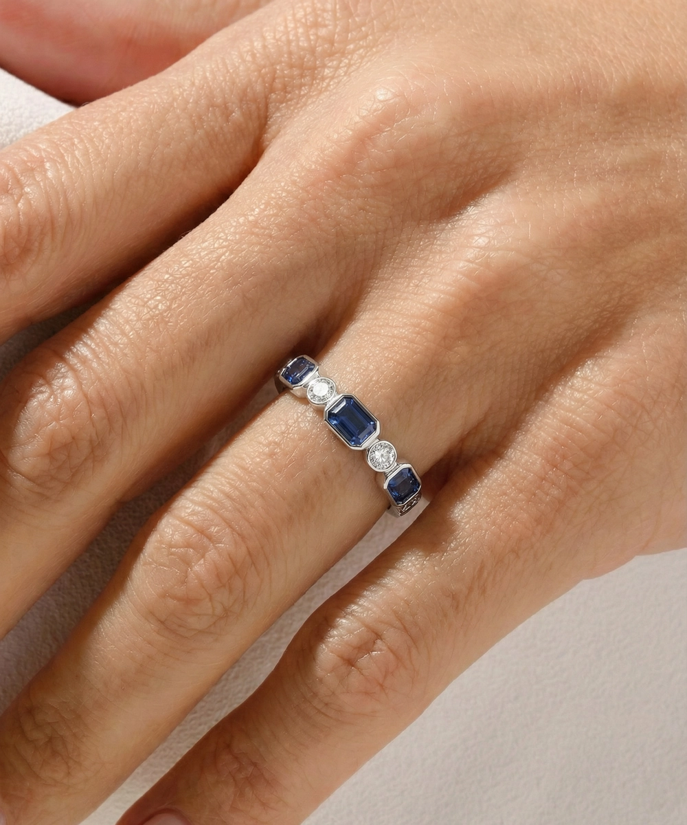 Close-up of a hand wearing a ring with blue gemstones and diamonds on a neutral background