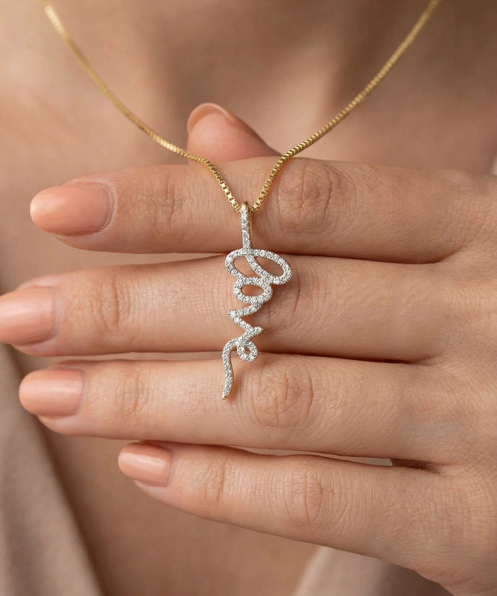 Gold necklace with a diamond pendant held by a hand against a neutral background