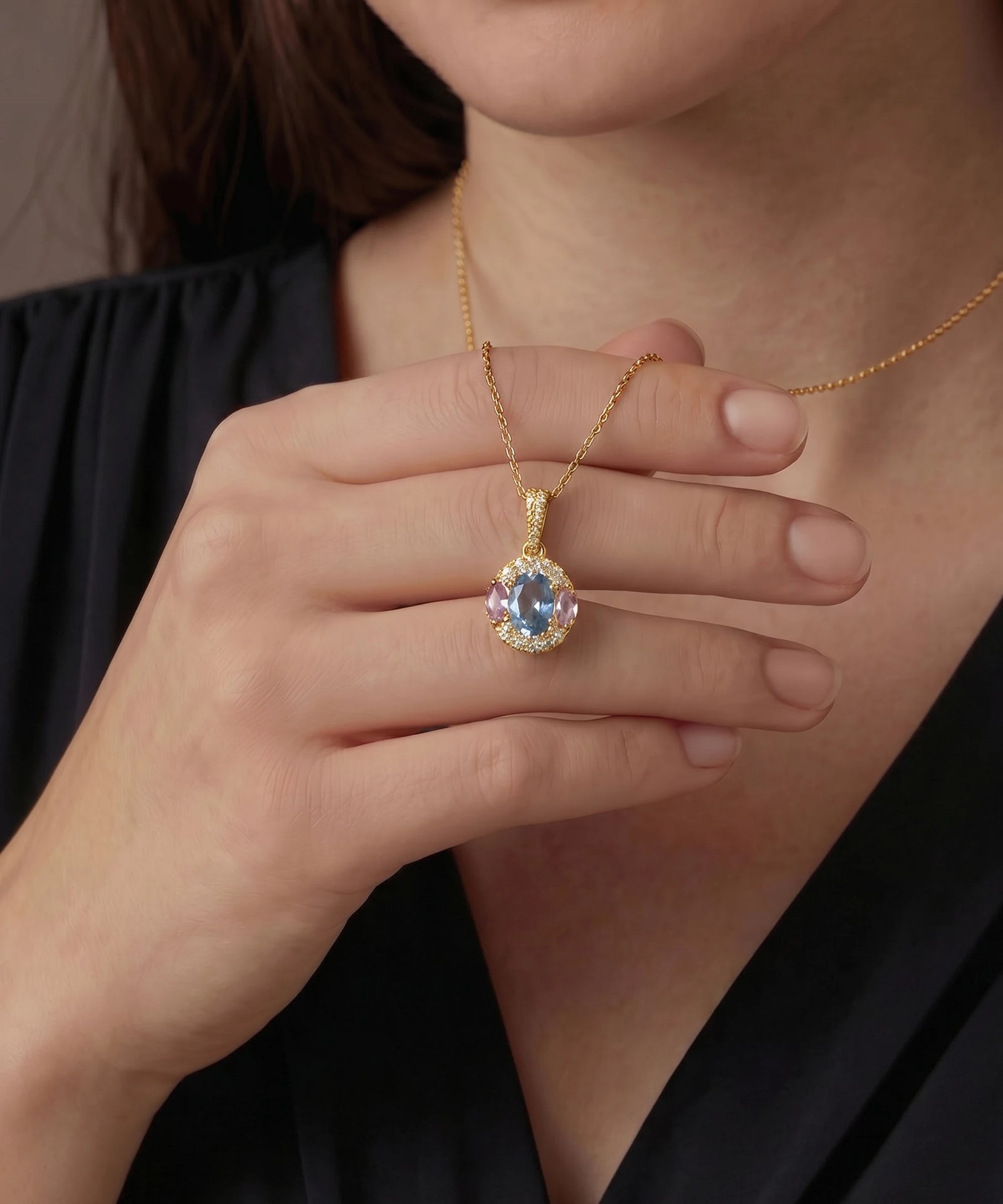 Gold necklace with a pendant featuring a blue gemstone held by a hand against a dark background