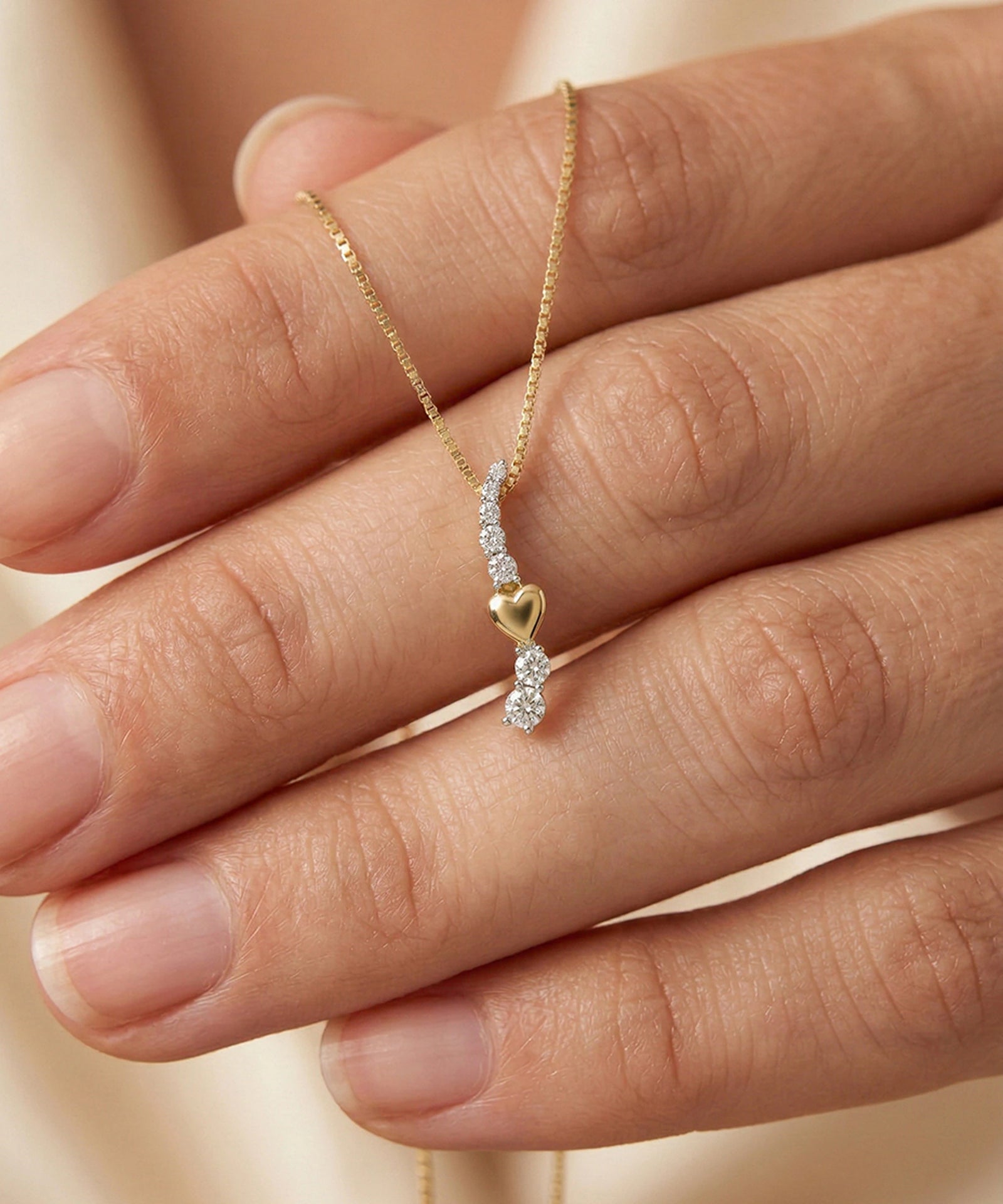 Gold necklace with a heart pendant held by a hand against a neutral background