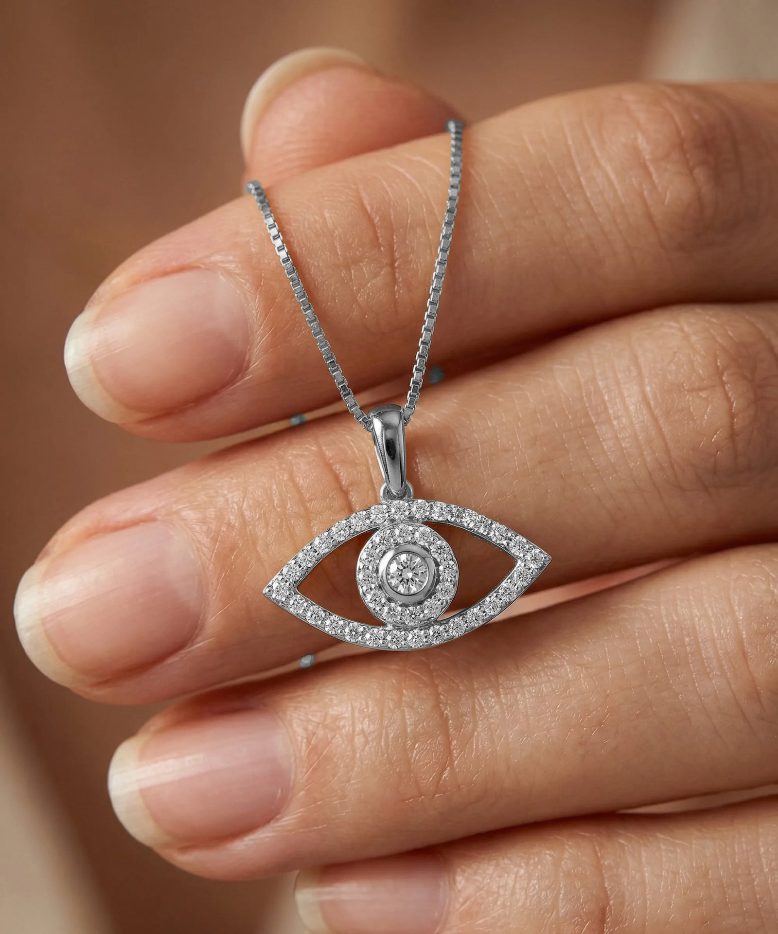 Silver eye-shaped pendant necklace held between fingers against a neutral background