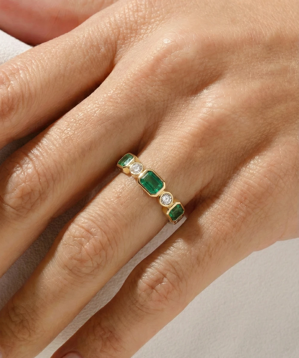 Gold ring with emerald and diamond stones on a hand against a neutral background