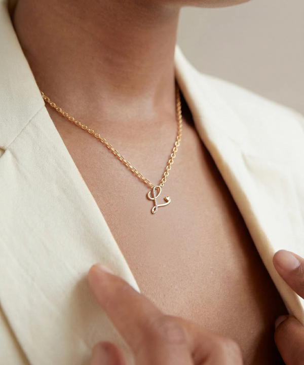 Gold necklace with a letter 'C' pendant worn by a person in a cream blazer.