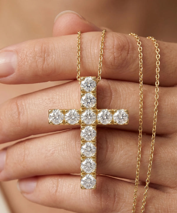 Gold cross necklace with diamonds held by a hand against a neutral background