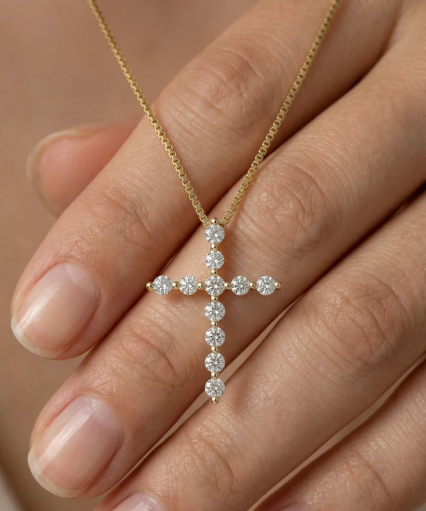Gold cross necklace with diamonds held between fingers against a neutral background