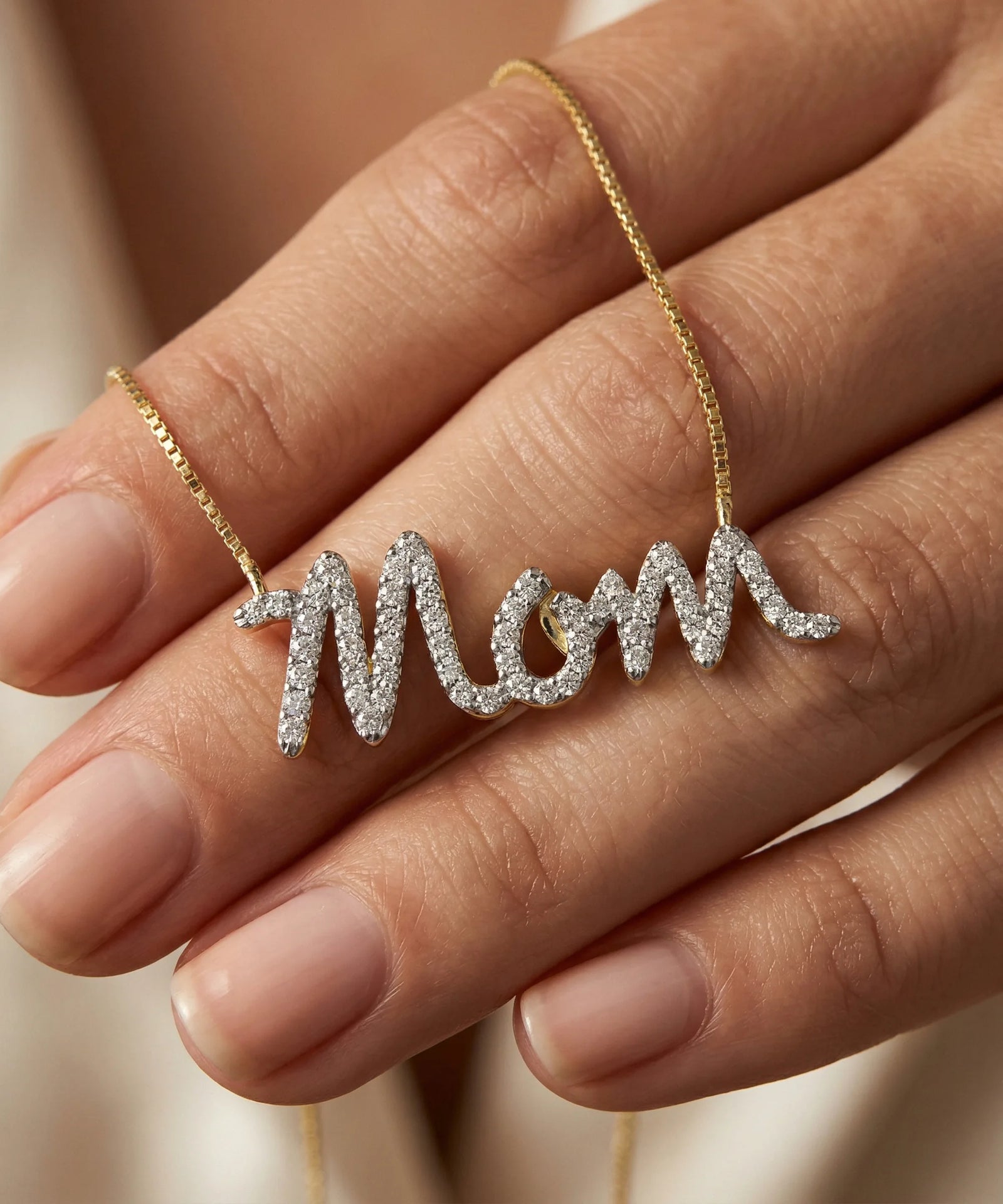 Gold necklace with 'Mom' pendant on a hand against a neutral background
