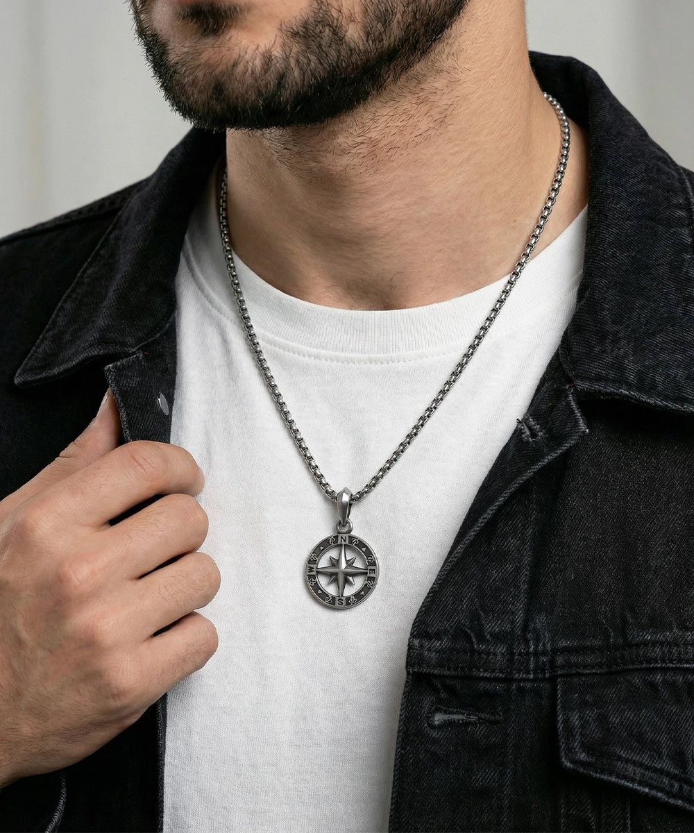Man wearing a black jacket and white shirt with a necklace featuring a compass pendant.