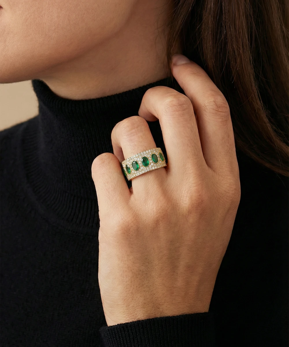 Close-up of a hand wearing a gold ring with green gemstones against a neutral background