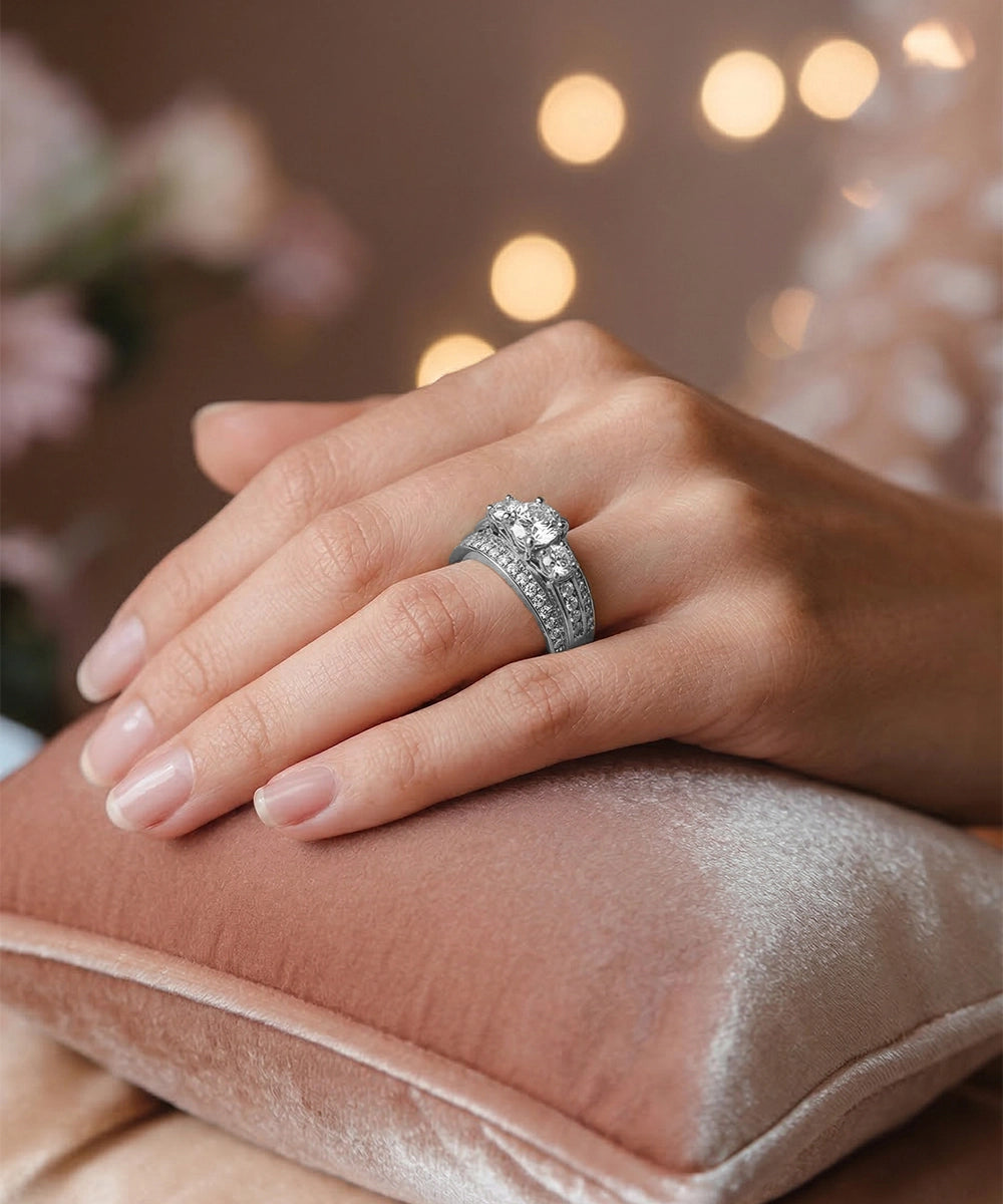 Hand wearing a diamond ring on a pink cushion with blurred lights in the background