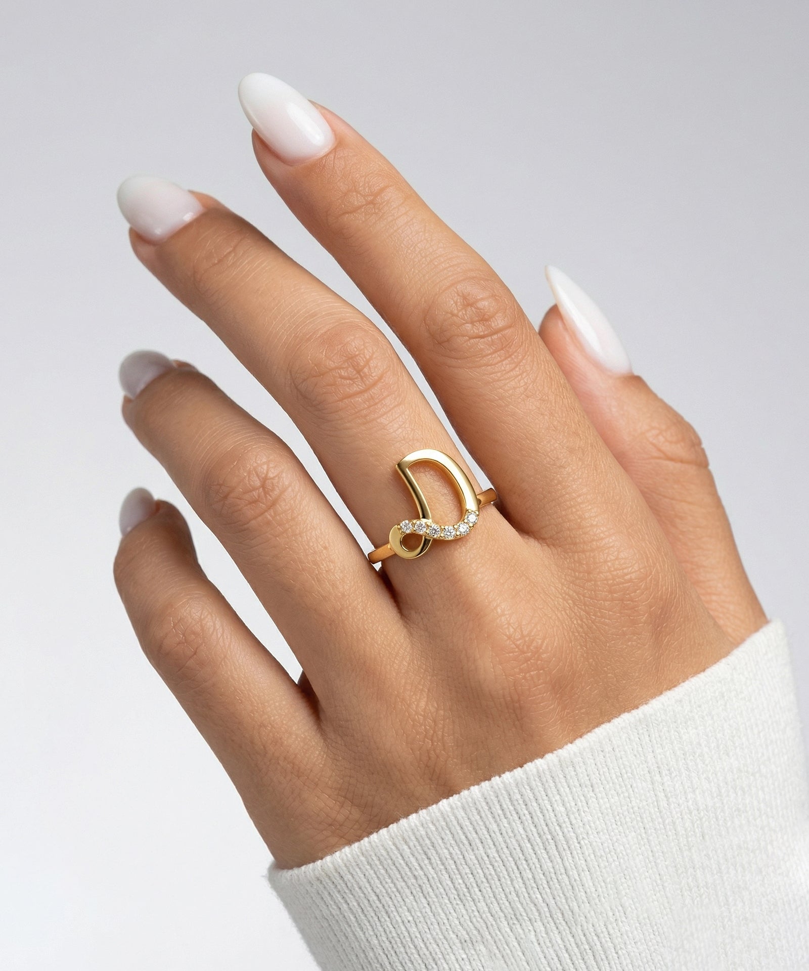 Gold ring on a hand with white nail polish against a light gray background