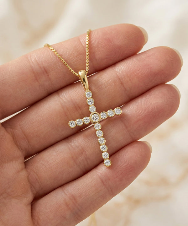 Gold cross-shaped necklace with clear stones held in a hand against a neutral background