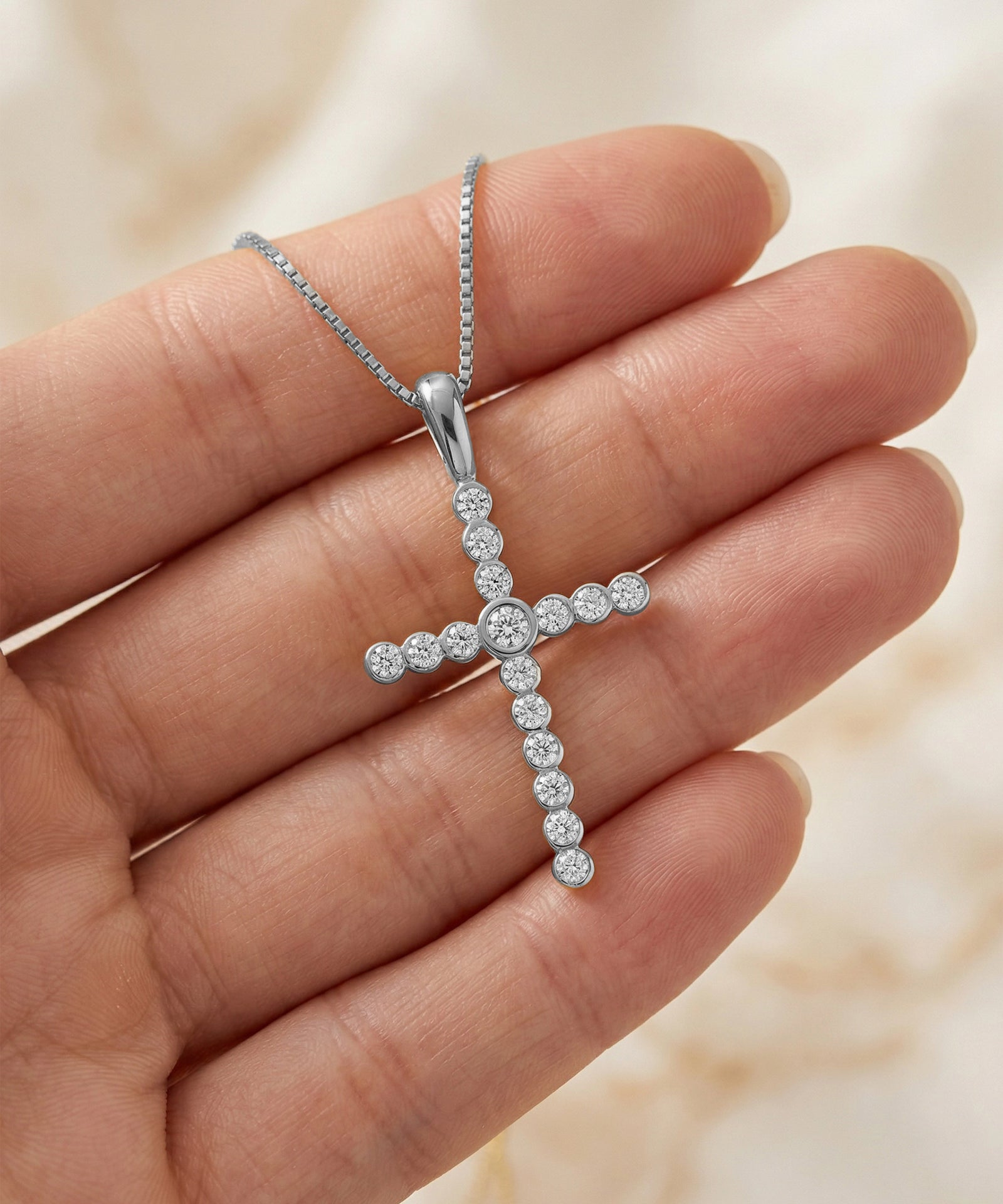 Silver cross pendant with diamonds held in a hand against a neutral background