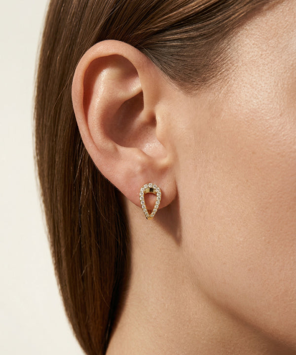 Close-up of an ear wearing a gold earring with a clear gemstone on a neutral background