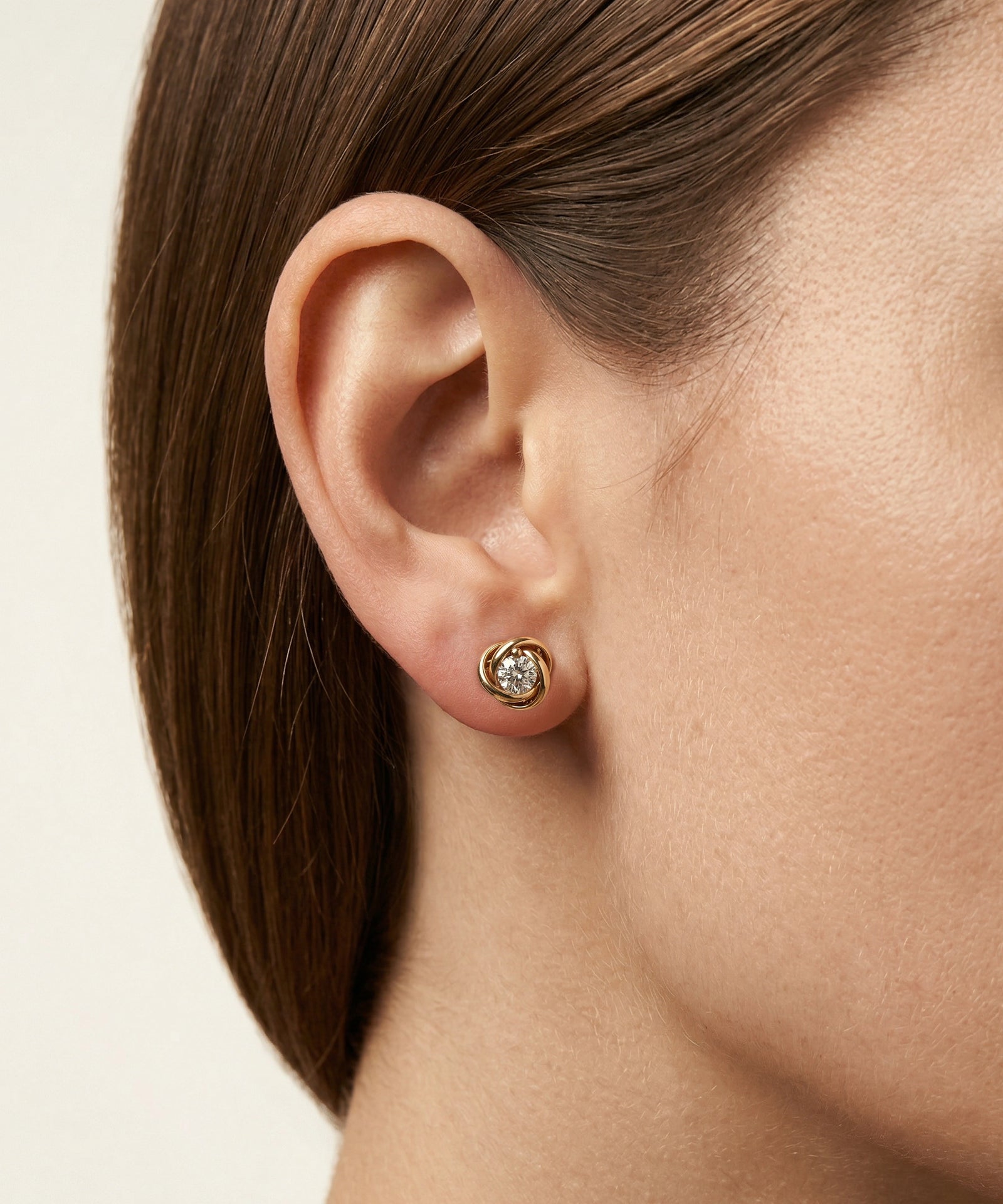 Close-up of an ear wearing a gold earring with a clear gemstone on a neutral background