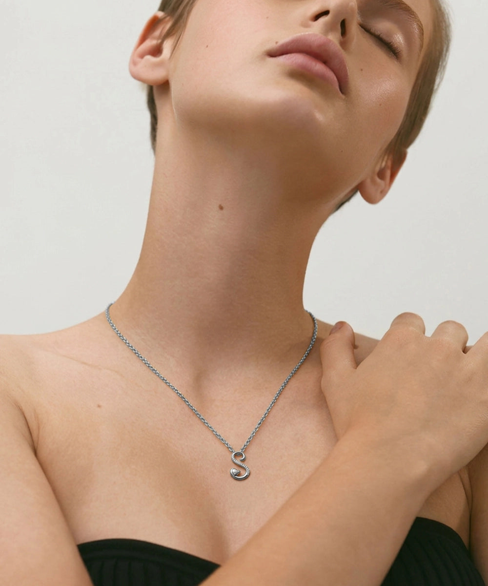 Person wearing a silver necklace with a 'S' pendant on a plain background