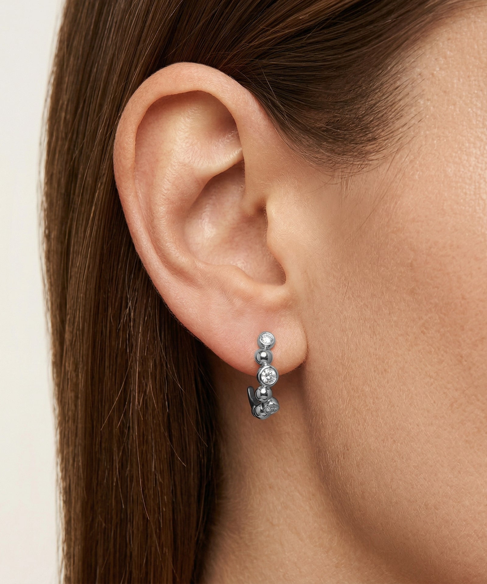 Close-up of an ear wearing a silver earring with three clear stones on a neutral background