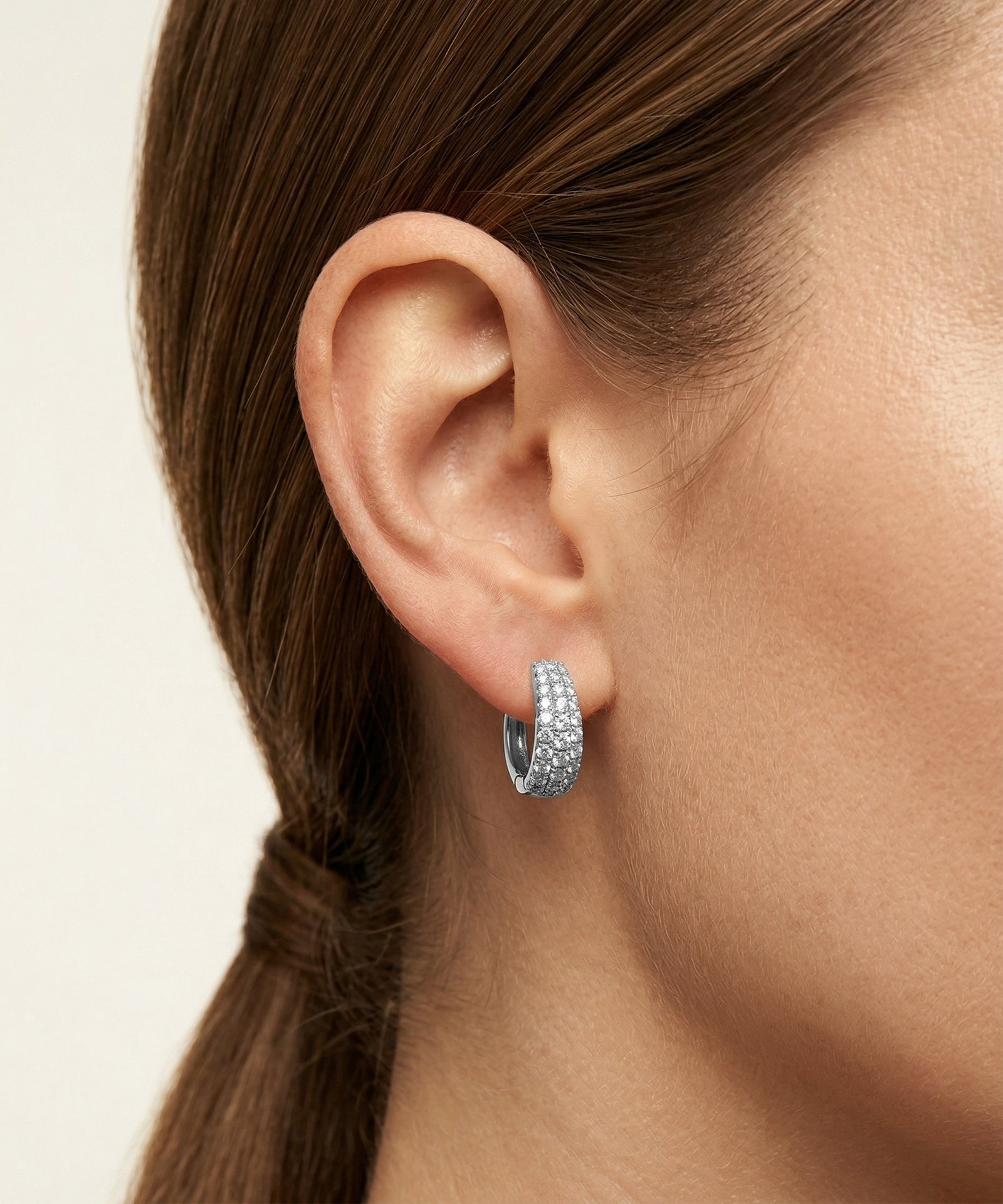 Close-up of a person wearing a silver hoop earring with clear stones on a neutral background