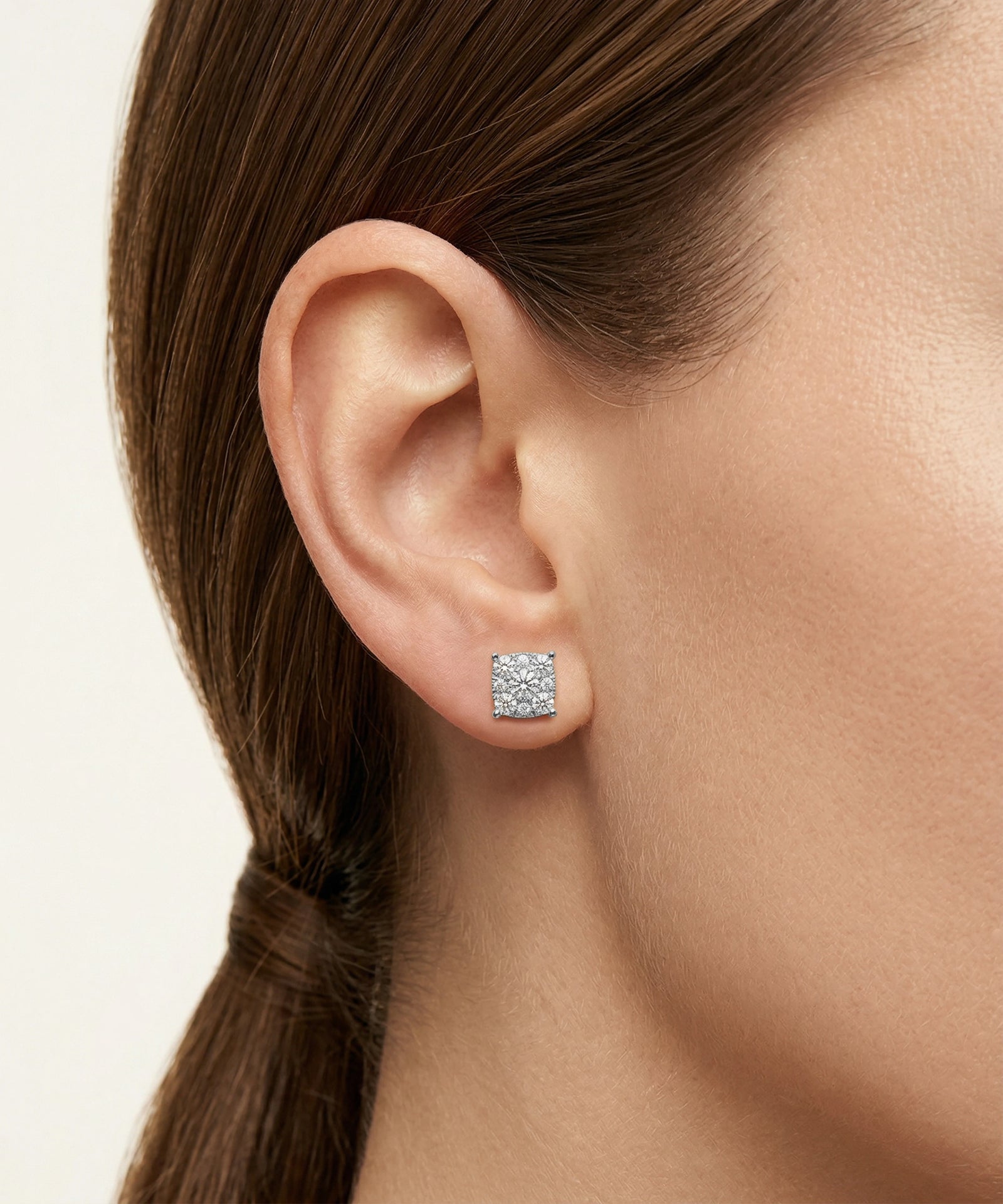 Close-up of an ear wearing a square stud earring with a clear gemstone on a neutral background