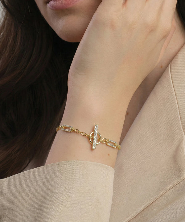 Gold bracelet with a pearl charm on a person's wrist, wearing a beige outfit.