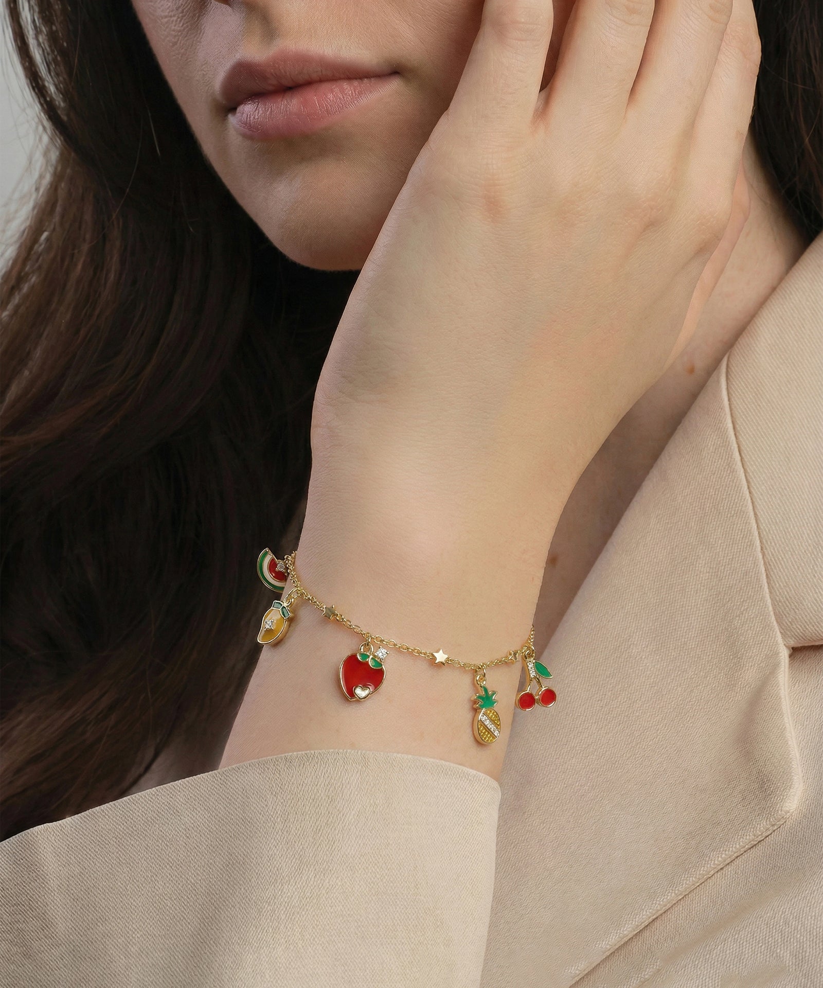 Close-up of a person wearing a gold bracelet with fruit charms on a neutral background