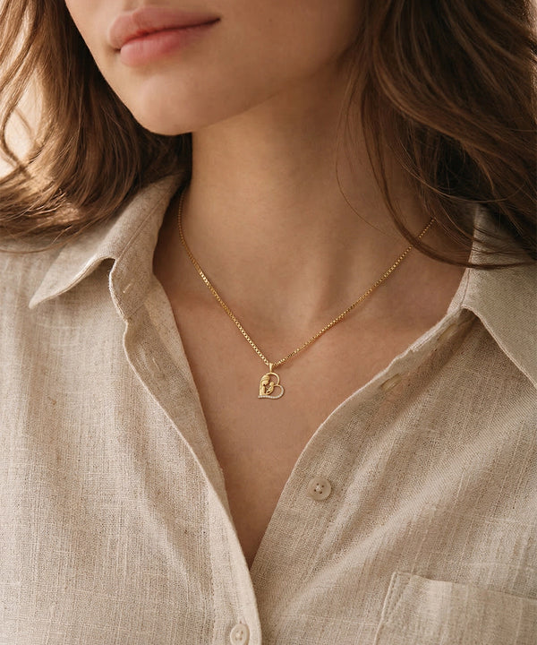 Close-up of a person wearing a gold necklace with a heart pendant, set against a neutral background.