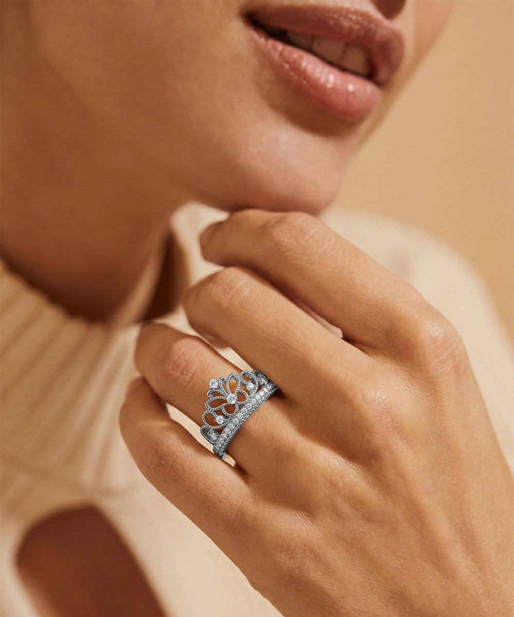 Close-up of a hand wearing a silver ring with a tiara design, against a soft-focus background.