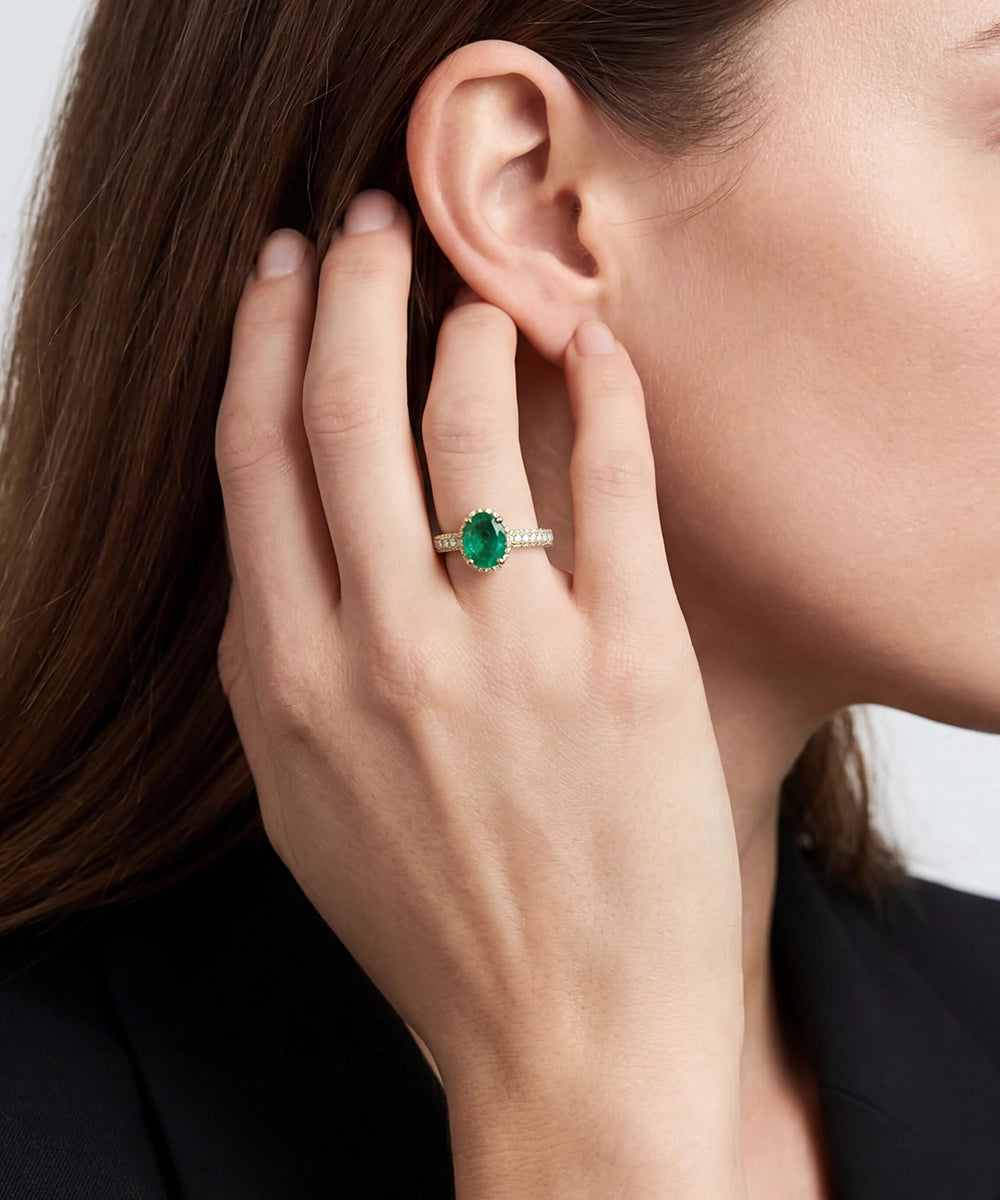 Close-up of a hand wearing an emerald ring, touching the ear against a neutral background
