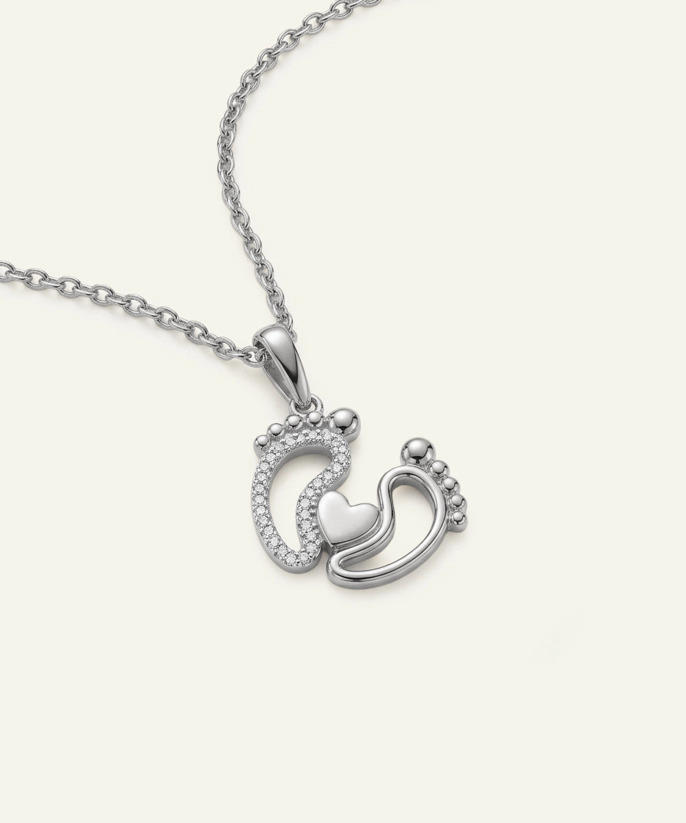 Silver necklace with a pendant featuring two interlocking footprints and a heart on a white background.