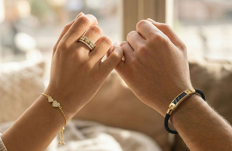 Couple holding pinky fingers wearing diamond ring and gold bracelets in warm indoor setting