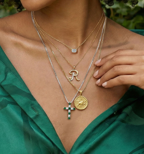 Woman wearing layered gold and silver necklaces with diamond pendant, initial “R” charm, cross, and coin medallion over green blouse